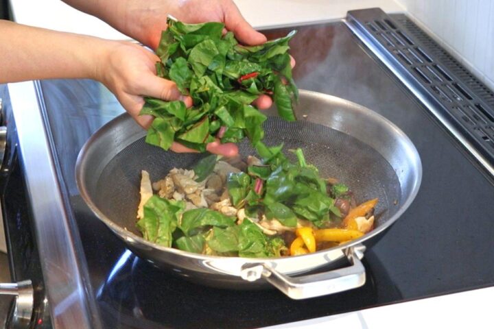 Someone adding chopped swiss chard into a wok of cooked vegetables and chicken.