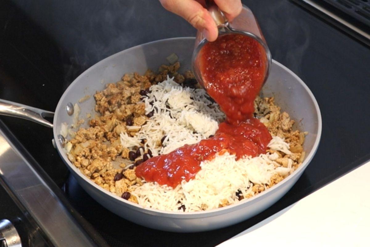 Someone adding salsa to a frying pan of ground turkey and rice.