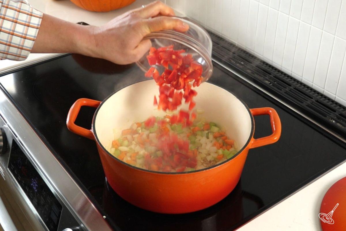 Someone adding chopped bell peppers to a large soup pot with other vegetables. 