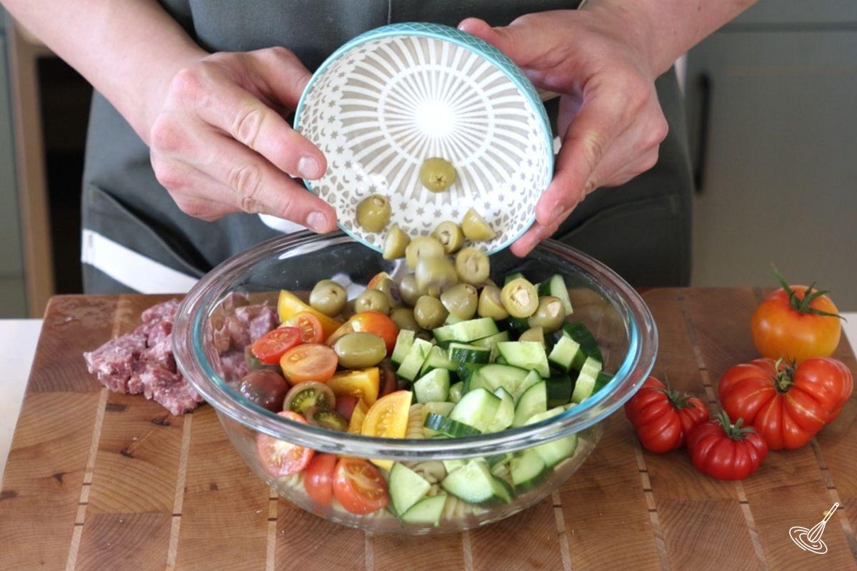 Someone adding sliced green olives on top of a bowl of cucumber tomato pasta salad.