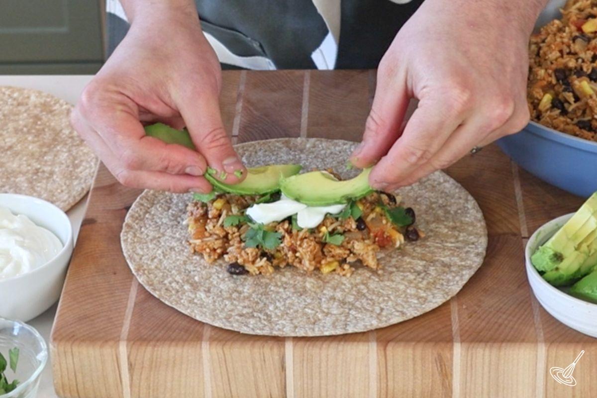 Someone placing slices of avocado on a flour tortilla with a rice and turkey mixture.