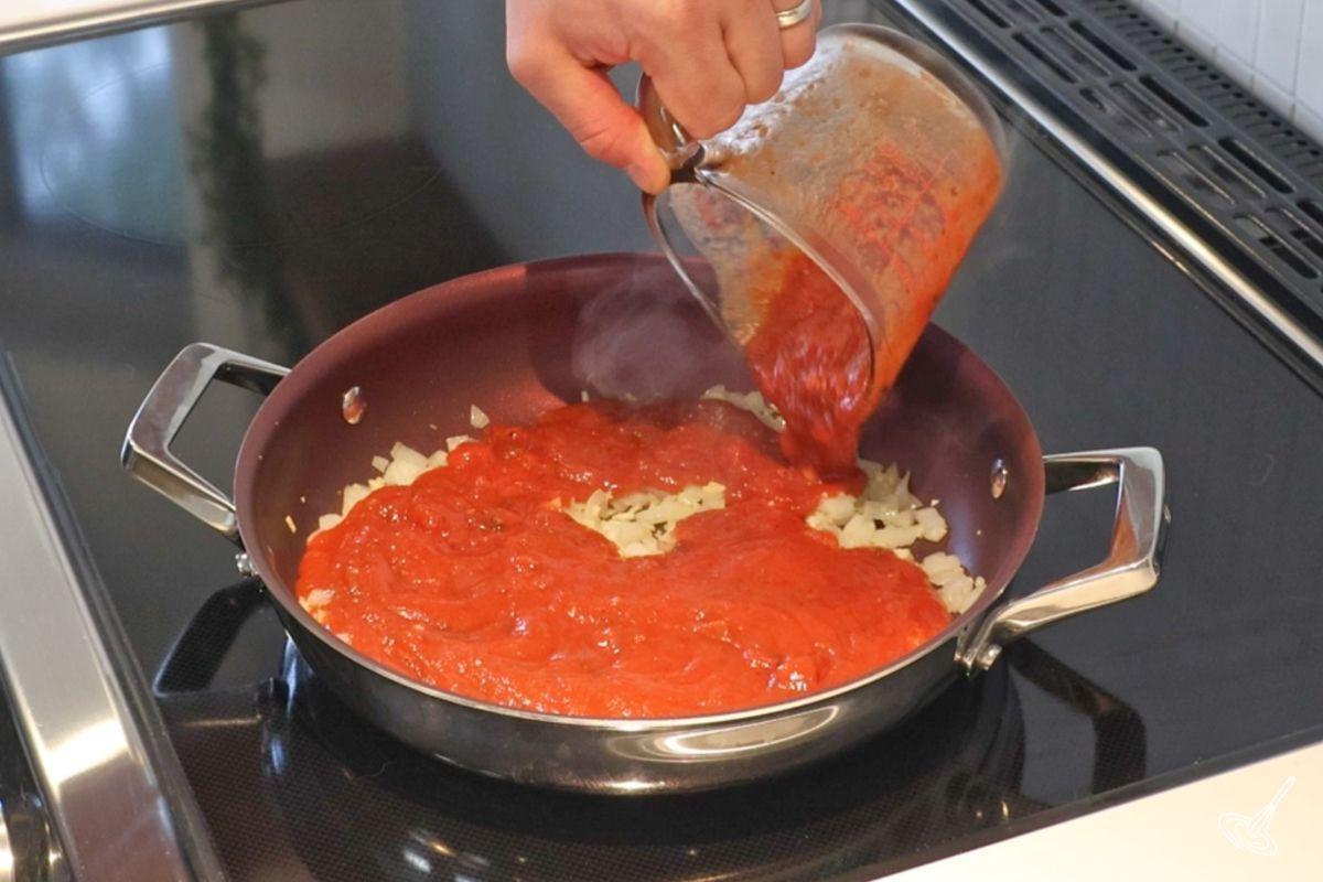 Someone adding marinara sauce to a large skillet.