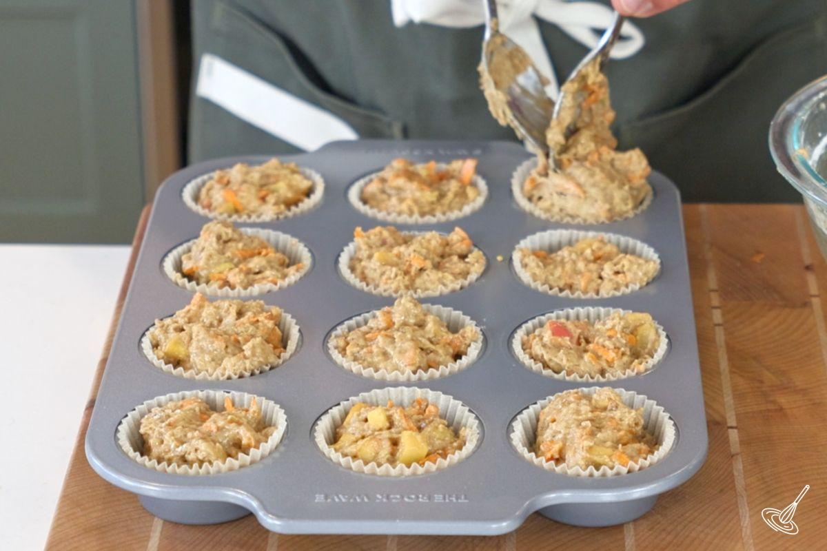 Someone using spoons to place muffin batter in a lined baking tray.