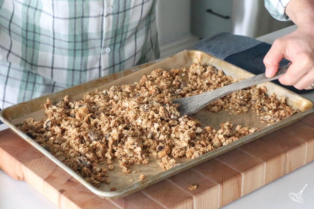 Someone using a wooden spoon to move around baking granola on a tray.