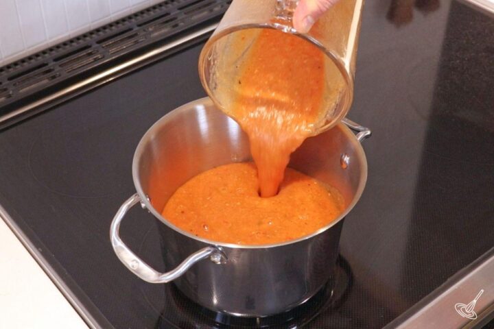 Someone pouring blended roasted tomato and garlic soup in a pot. 