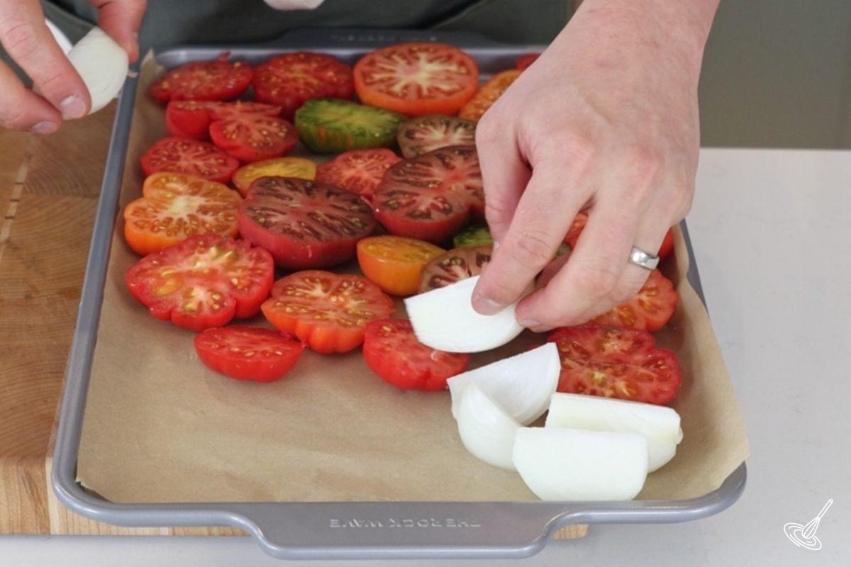 Someone placing pieces of onion a baking tray with halved tomatoes.
