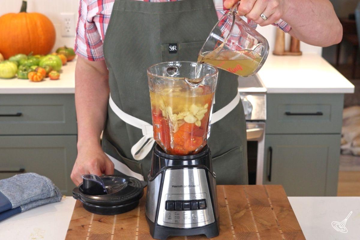 Someone adding chicken broth to a blender with roasted tomatoes and garlic. 