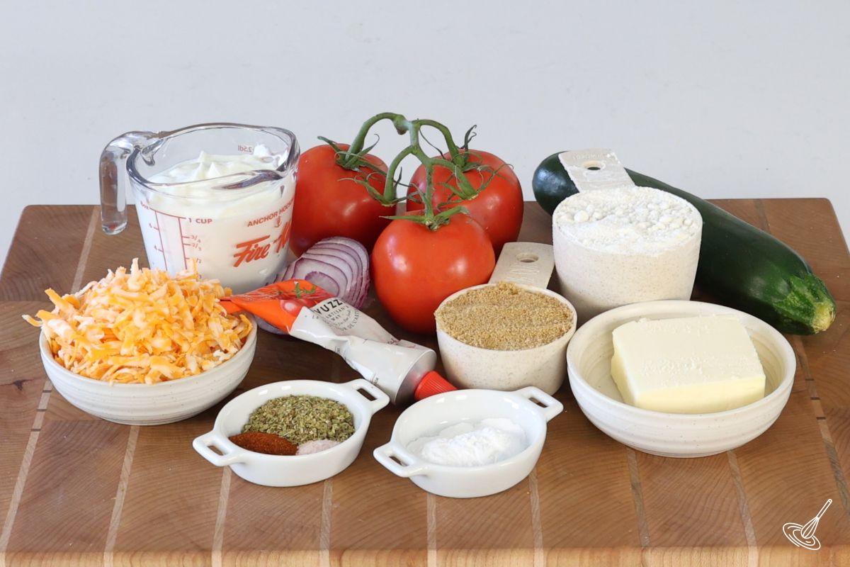 Ingrédients disposés sur une planche à découper, comprenant du fromage, du yogourt, du beurre, de la farine, des tomates et des courgettes.