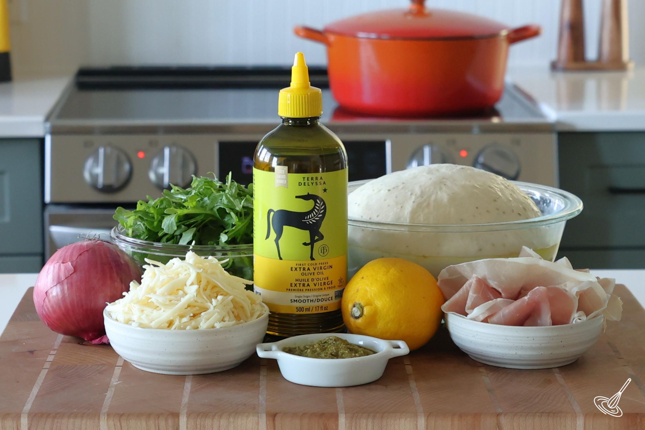 Ingredients to make a Prosciutto Arugula Pizza, including pizza dough, olive oil, grated cheese, arugula, and prosciutto.