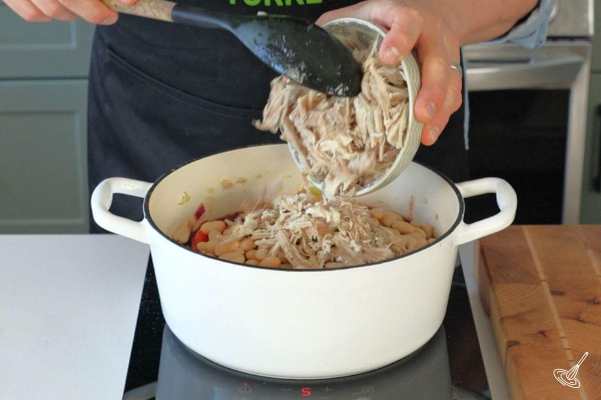 Someone adding shredded turkey to a Dutch oven of Cranberry Turkey chili. 