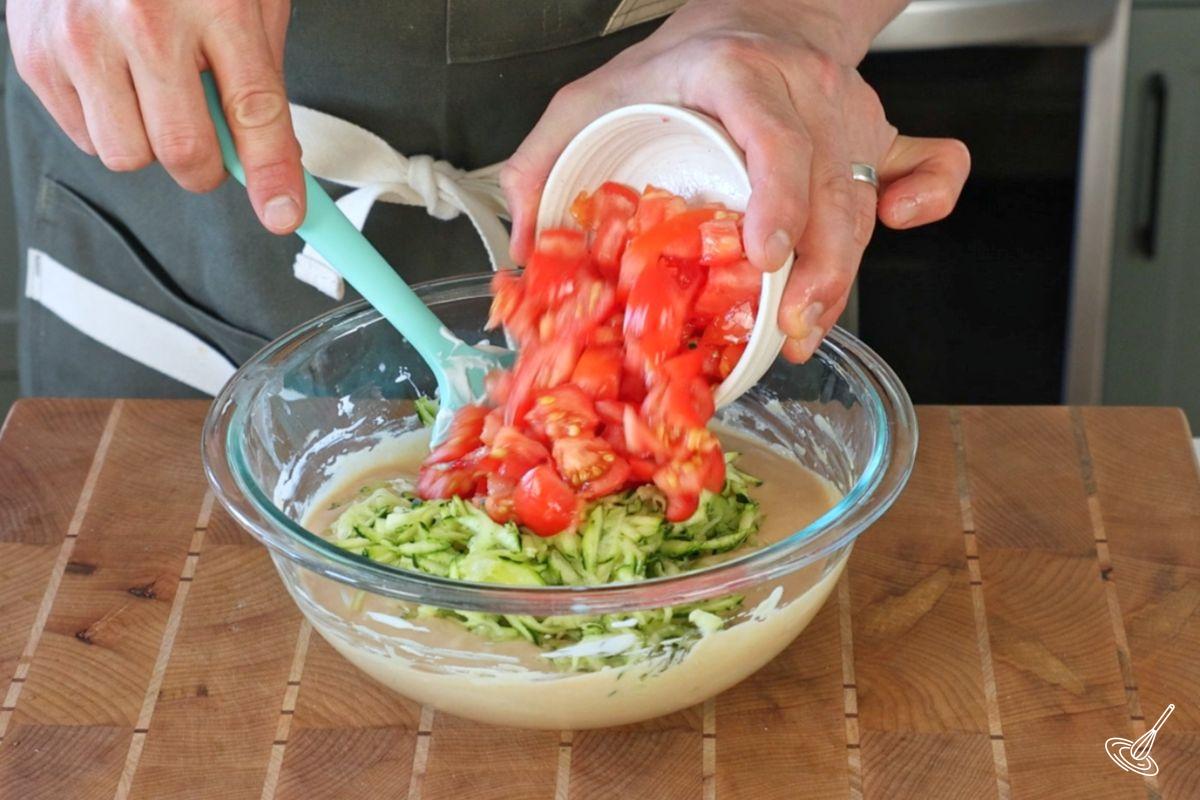 Quelqu’un ajoute des tomates en dés et de la courgette râpée dans un bol de pâte à muffins.