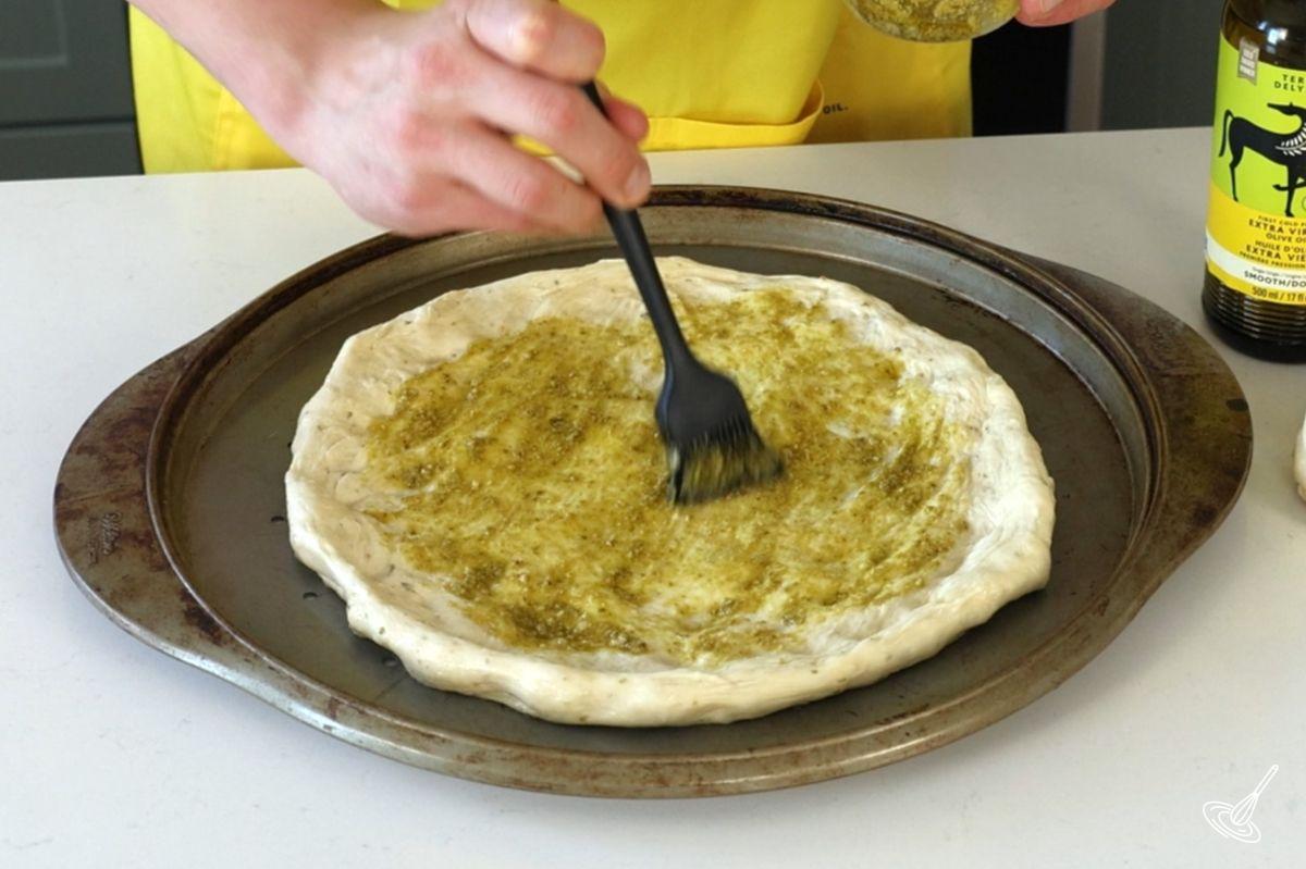 Someone using a silicone brush to spread pesto on a pizza crust. 