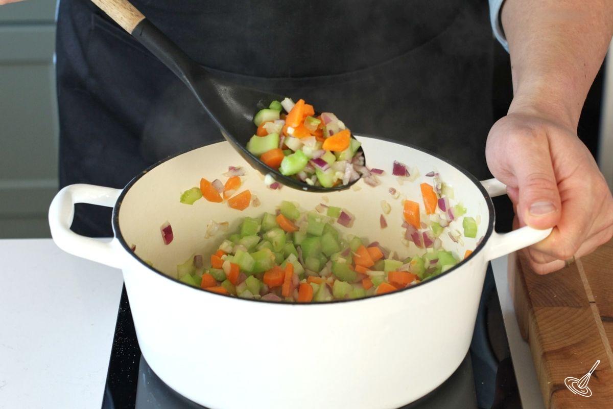Someone holding up a spoon of cooked diced vegetables over a Dutch oven. 