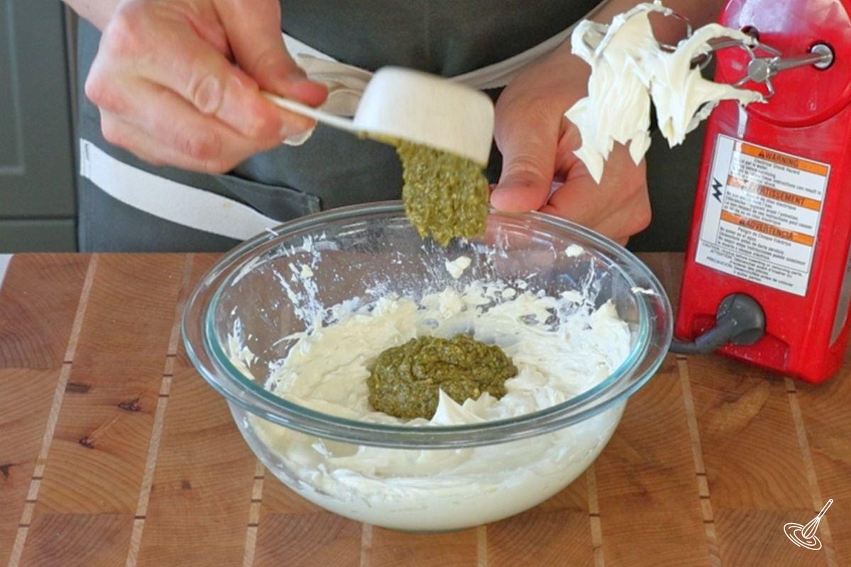 Quelqu’un ajoutant du pesto au basilic dans un bol de fromage à la crème. 