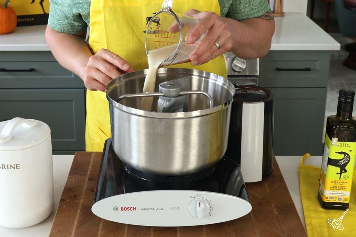 Someone pouring a yeast mixture in a mixer to make pizza dough. 