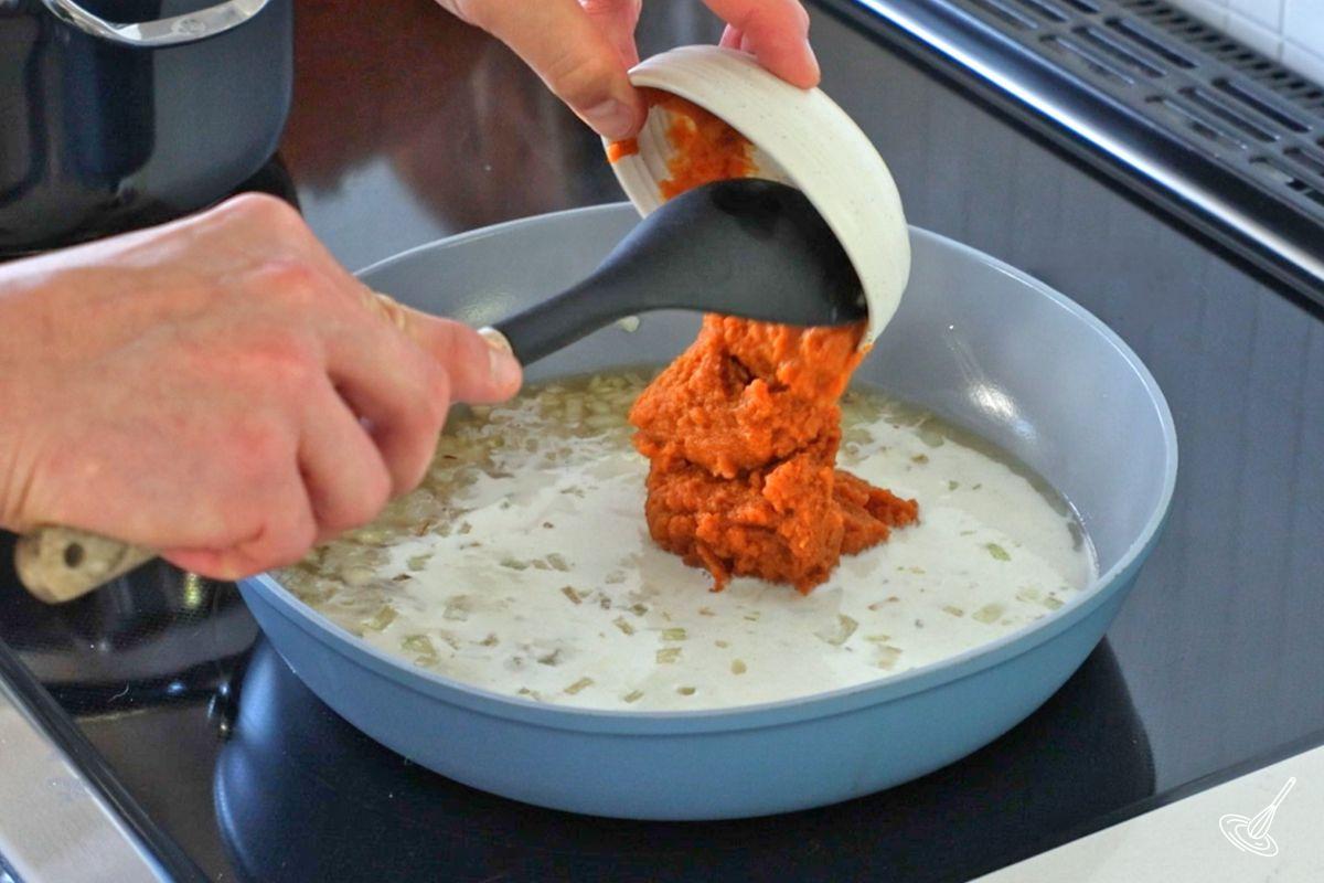 Someone adding pumpkin puree to a frying pan with chicken broth and cream.
