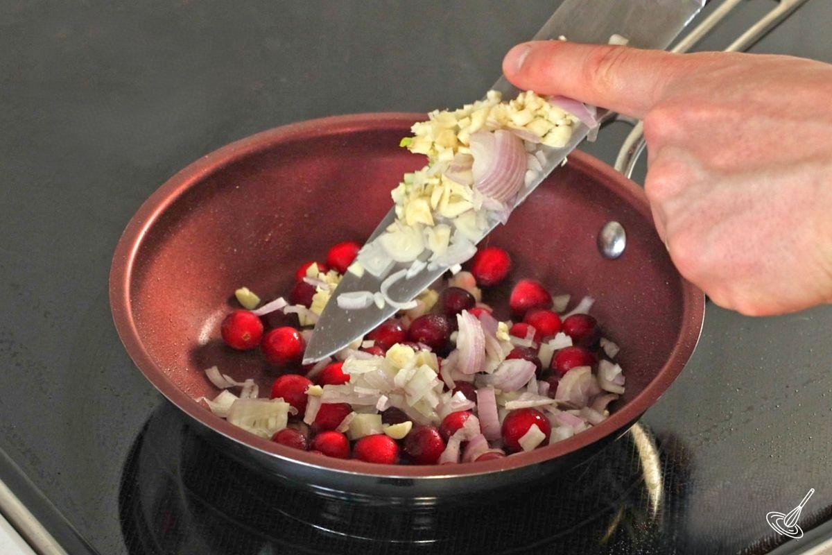 Someone adding chopped shallot and garlic to a frying pan with cranberries. 