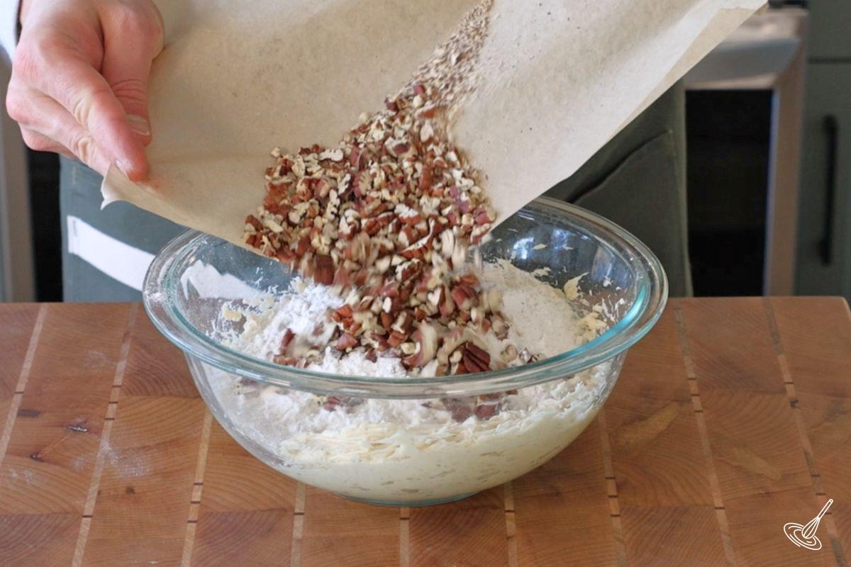 Someone adding toasted pecan pieces to a bowl of shortbread batter.