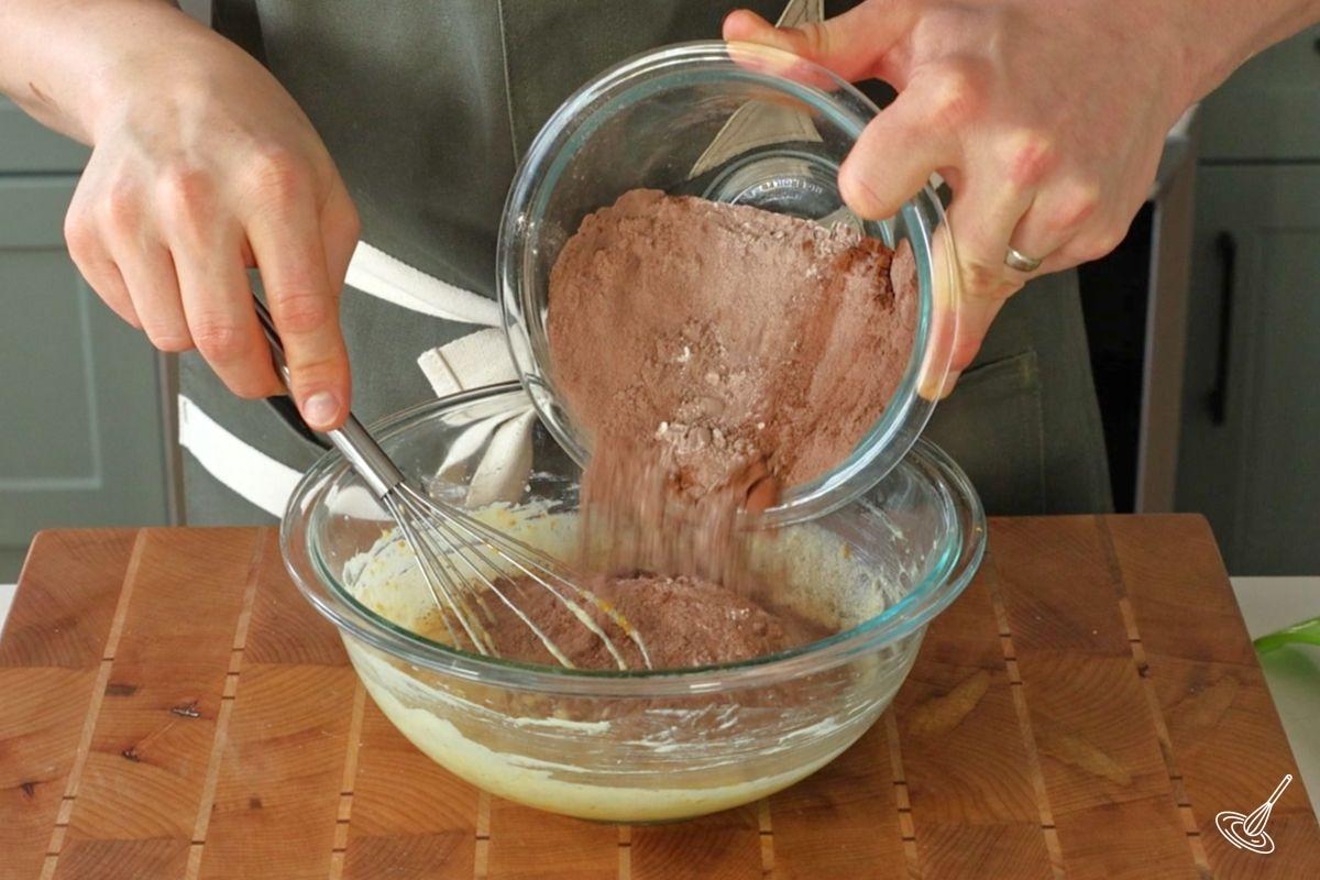 Someone adding dry ingredients to a bowl of batter.