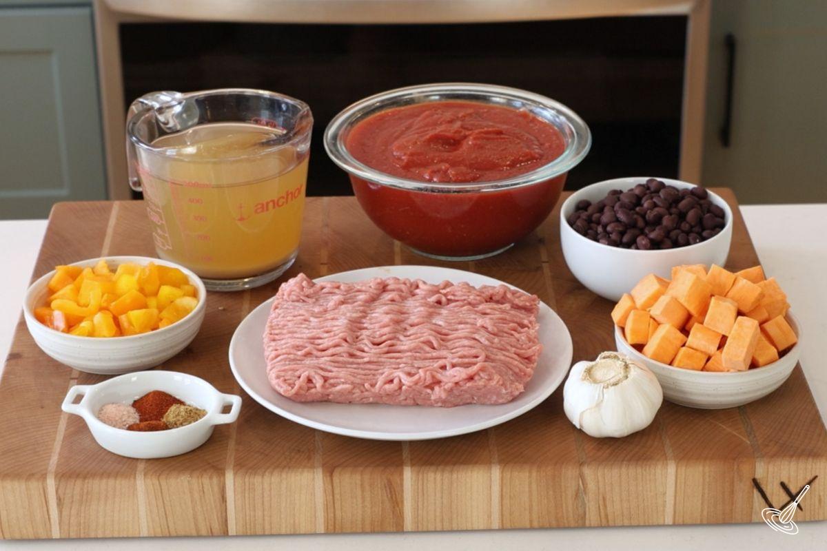 Ingrédients pour préparer le chili à la dinde et à la patate douce sur une planche à découper, incluant de la dinde hachée, des tomates écrasées, des patates douces et des haricots noirs.