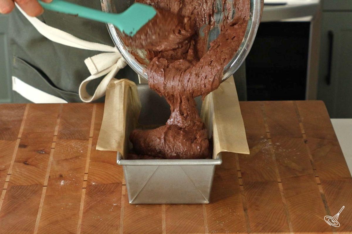 Someone pouring chocolate cake batter in a loaf pan.