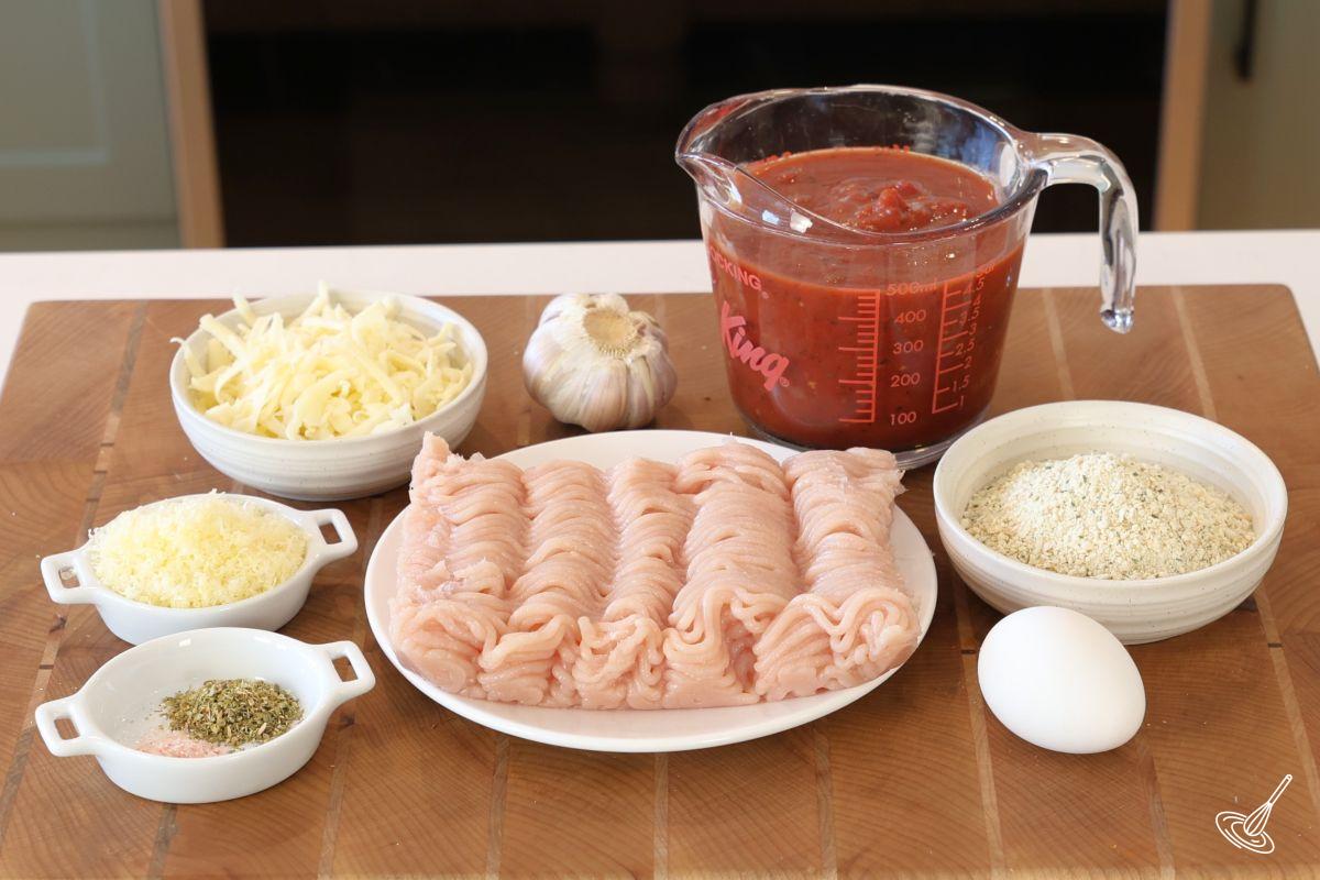 Ingredients on a wooden cutting board including; ground chicken, bread crumbs, tomato sauce, and cheese.