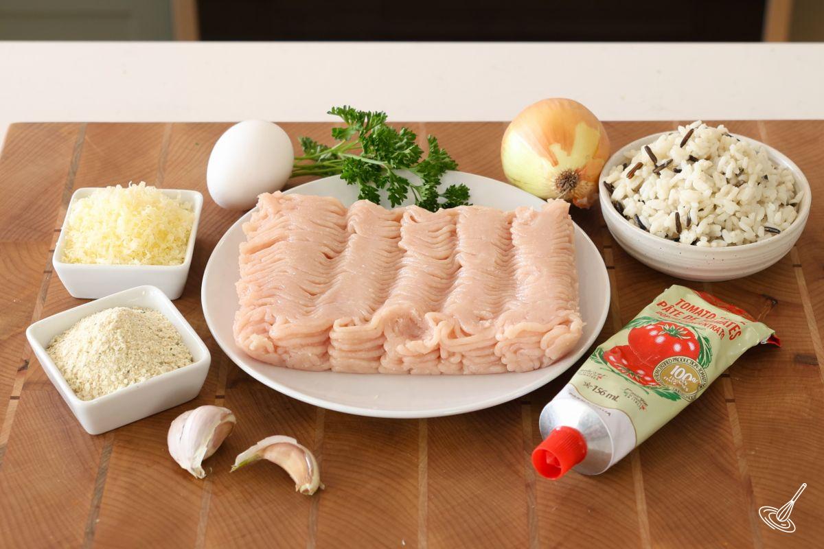Ingrédients disposés sur une planche à découper en bois : poulet haché, œuf, oignon, riz et chapelure.