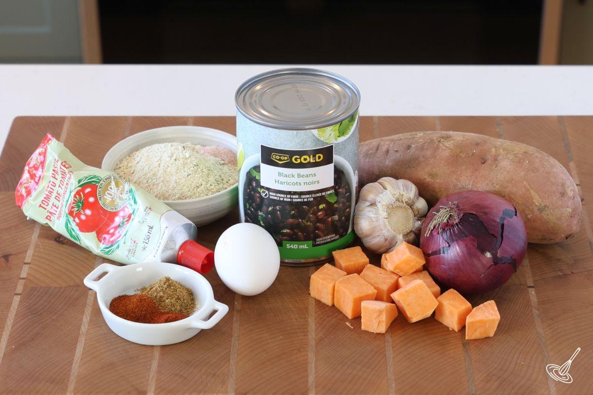 Ingrédients disposés sur une planche à découper en bois : une boîte de haricots noirs, une patate douce, de la chapelure et un œuf. 