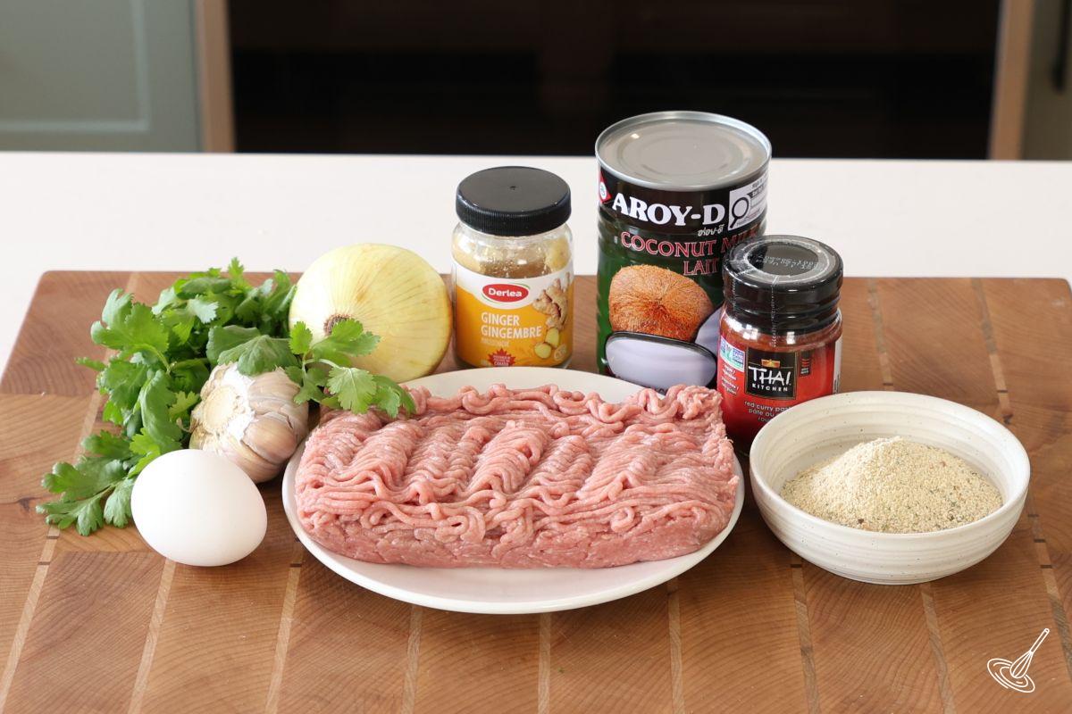 Ingredients on a cutting board to make meatballs, including, ground turkey, coconut milk, ginger, onion, garlic, egg, and breadcrumbs. 
