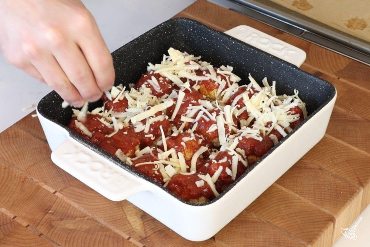 Someone adding grated mozzarella over a dish of chicken parm meatballs.