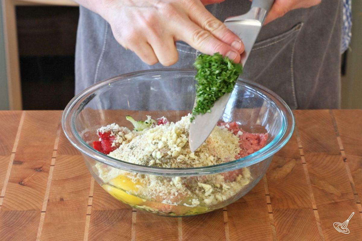 Quelqu’un ajoute du persil frais haché à un bol d’ingrédients pour préparer des boulettes de dinde et de courgette cuites au four. 
