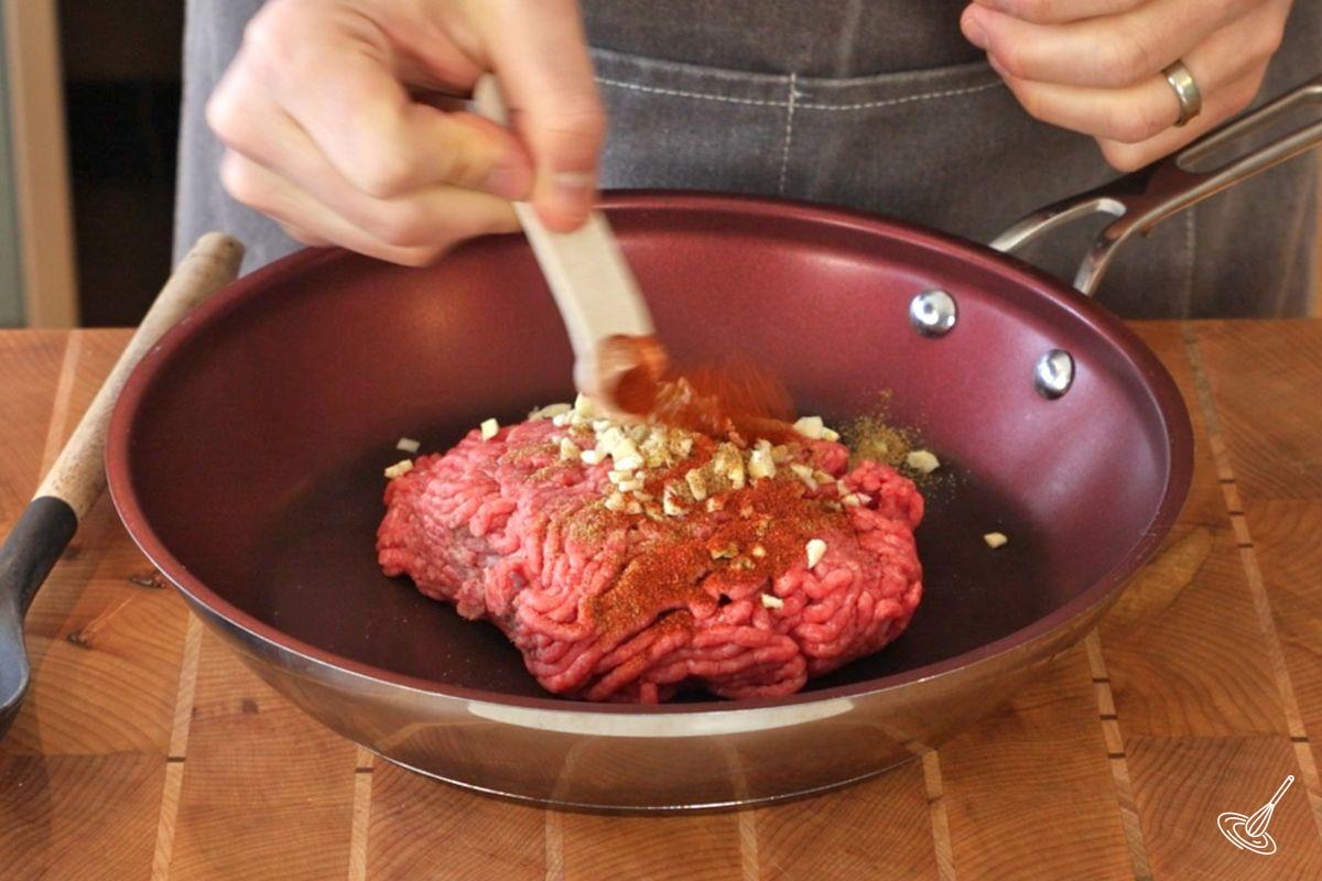 Someone adding seasoning to a frying pan of ground beef.