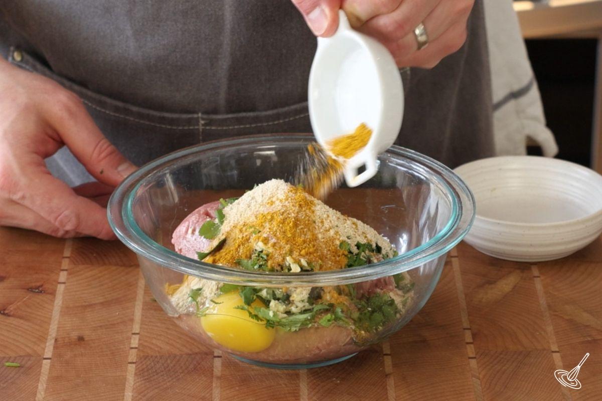 Someone adding curry powder to a bowl of meatball ingredients.