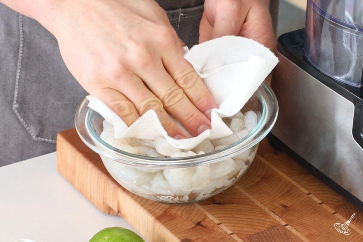 Someone using a paper towel to pat dry the raw shrimp.