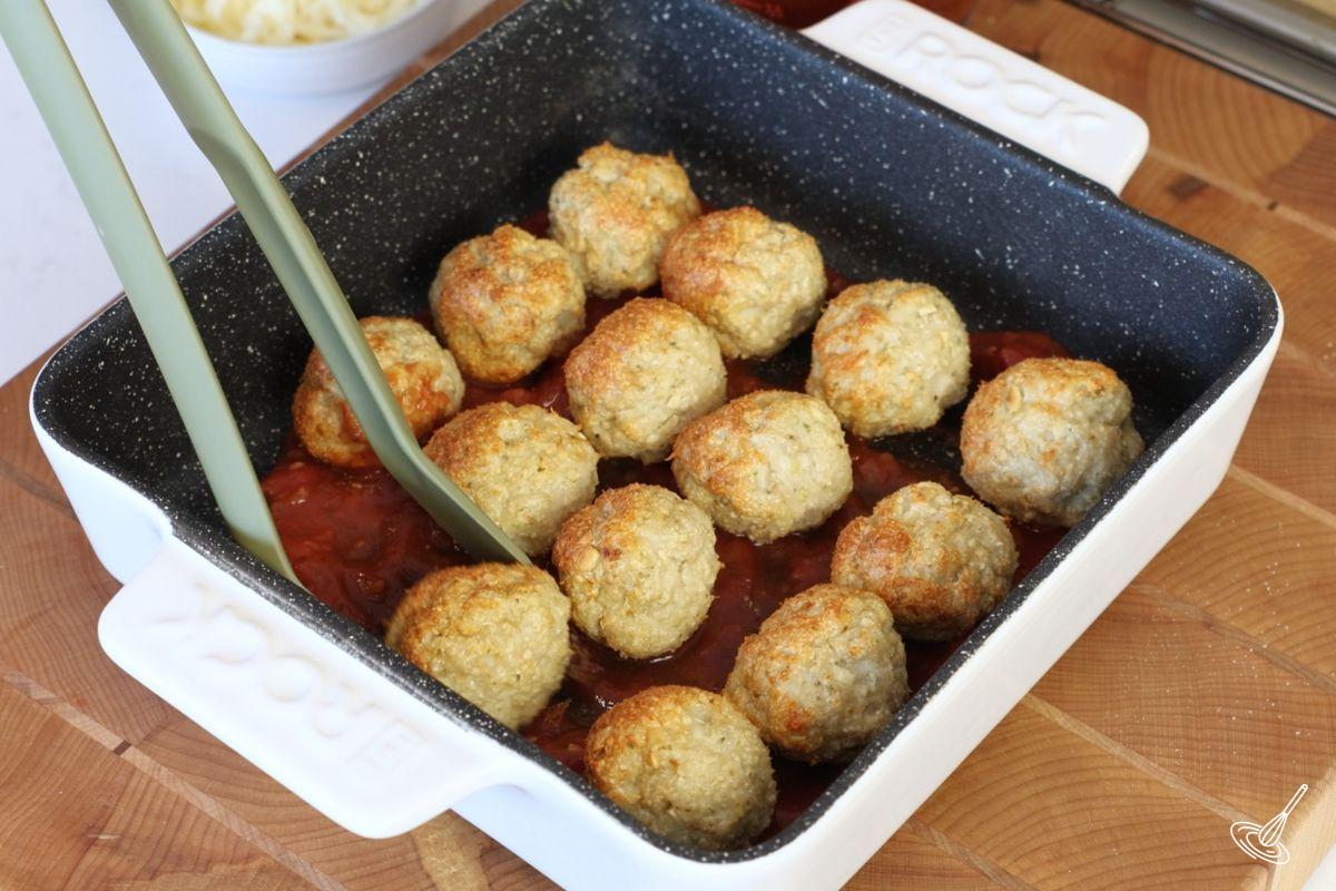 Someone placing cooked chicken parm meatballs in a baking dish with tomato sauce.