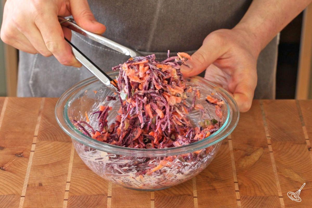 Someone using a pair of tongs to toss coleslaw into the buffalo dressing.