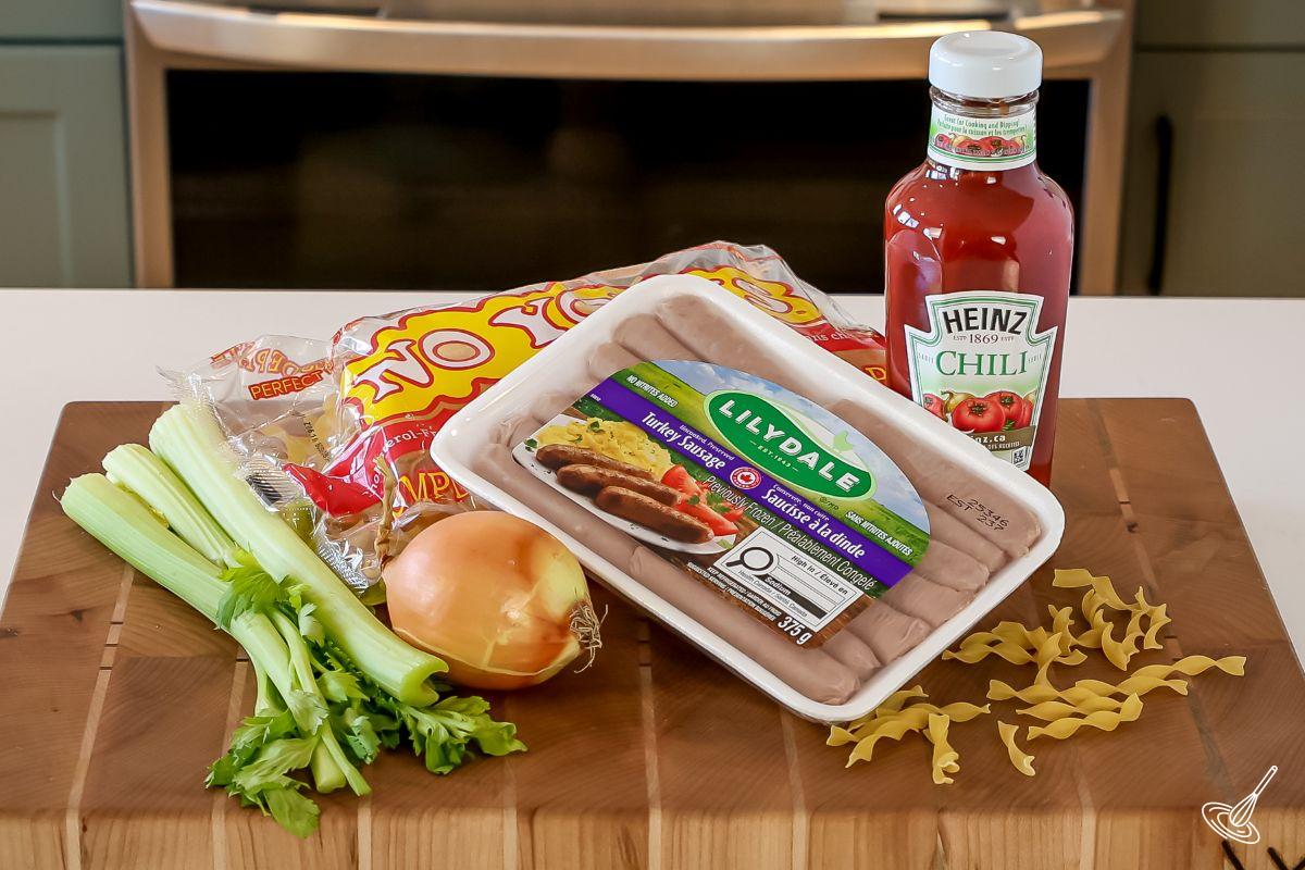 Ingrédients disposés sur une planche à découper, notamment des saucisses de dinde, du céleri, un oignon, de la sauce chili et des nouilles aux œufs.