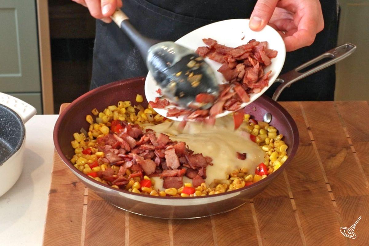 Someone adding crispy bacon pieces to a large pan of roasted corn kernels. 
