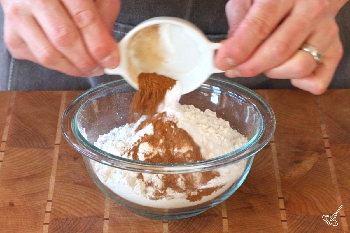 Someone adding cinnamon to a bowl of flour.