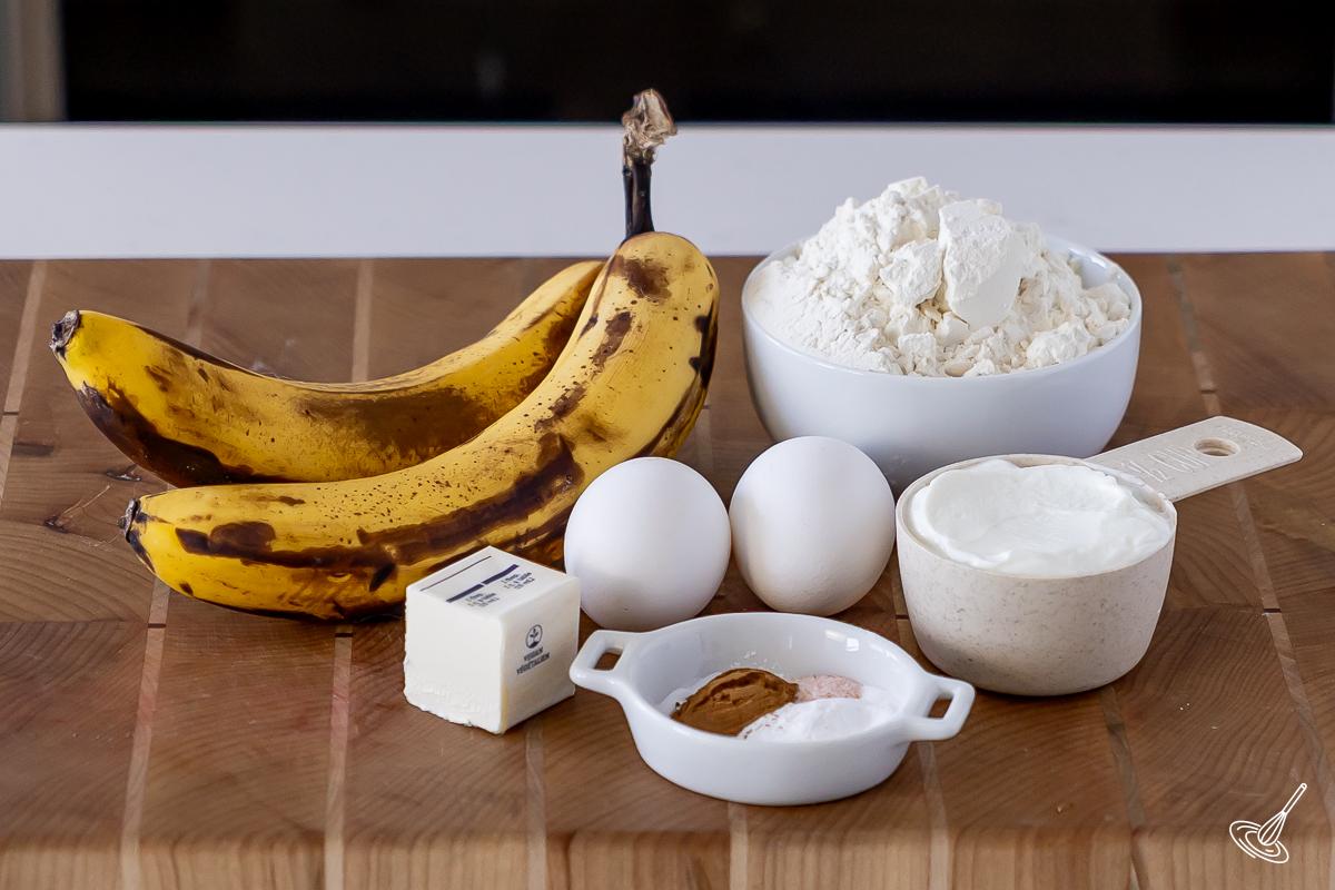 Ingrédients disposés sur une planche à découper, notamment des bananes, de la farine, des œufs et du yaourt grec.