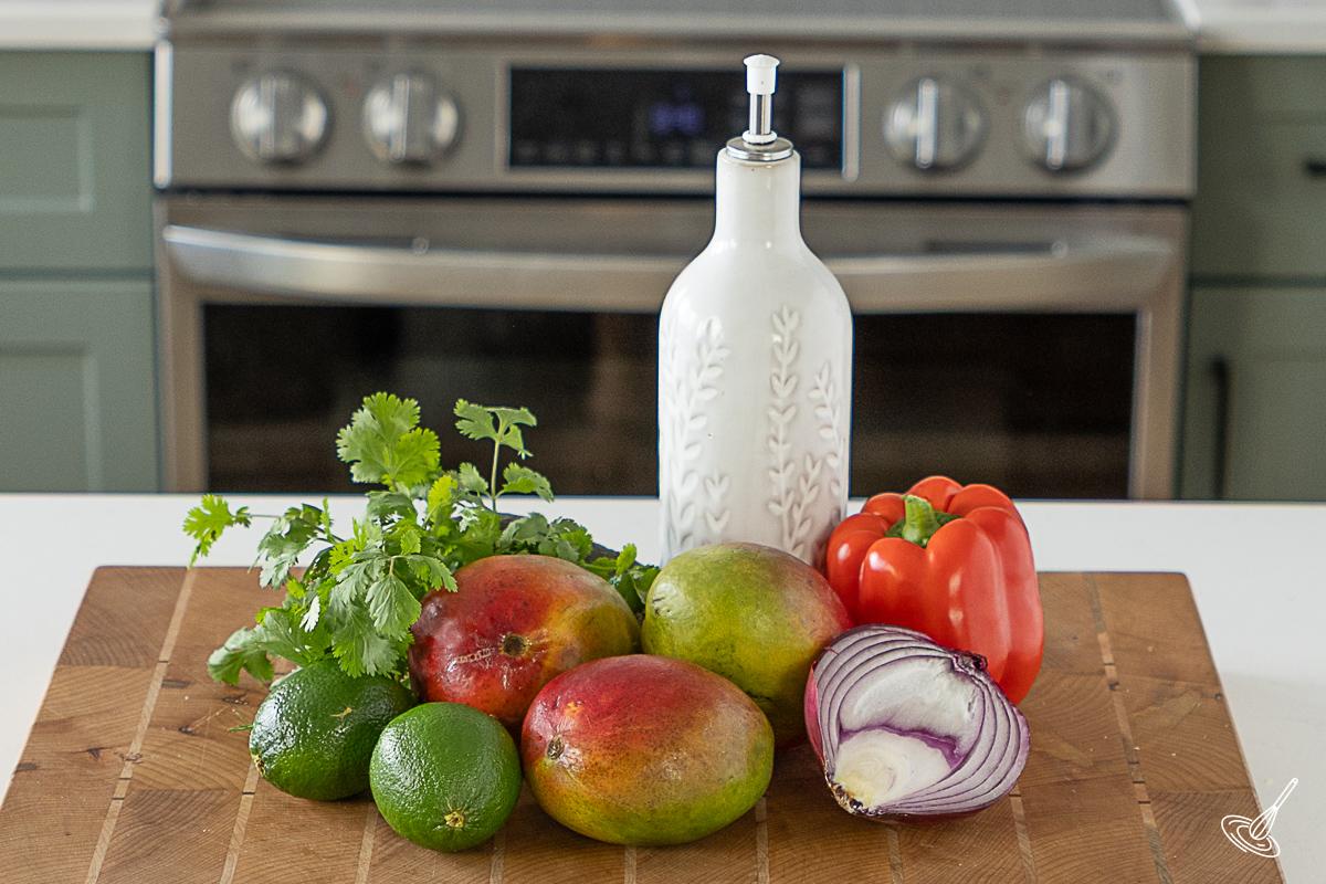 Ingredients on a cutting board to make Easy Mango Lime Salsa including mangos, limes, onion, and red bell pepper. 