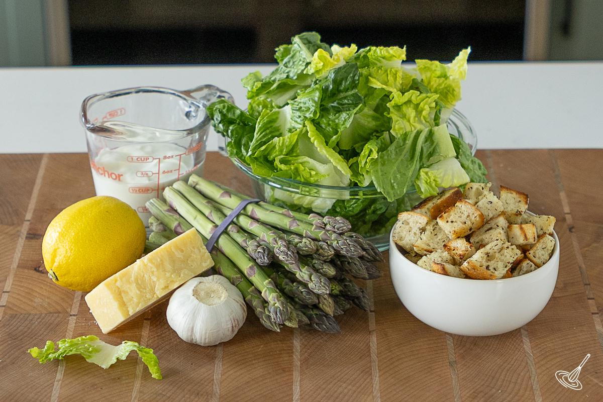Ingredients on a cutting board including chopped lettuce, a bunch of asparagus, croutons, and salad dressing.