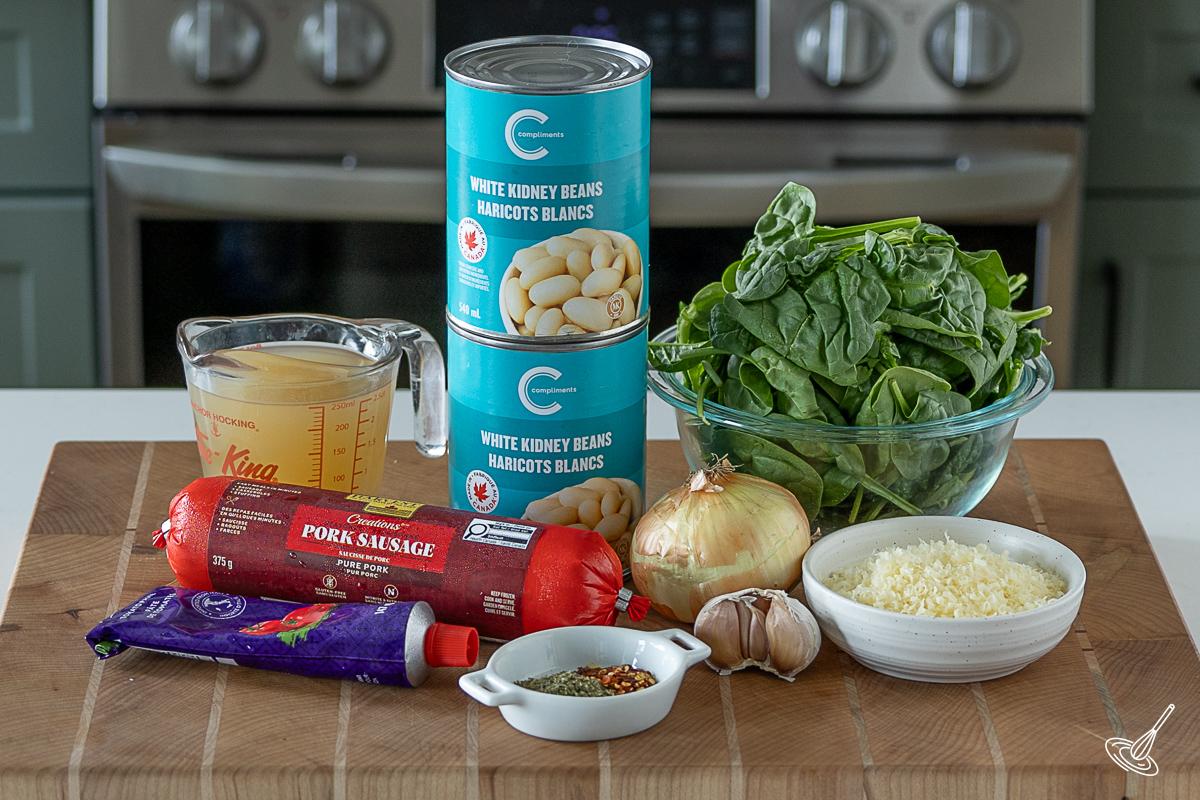 Ingredients on a cutting board including fresh spinach, pork sausage, cans of white kidney beans, chicken broth, and parmesan cheese. 
