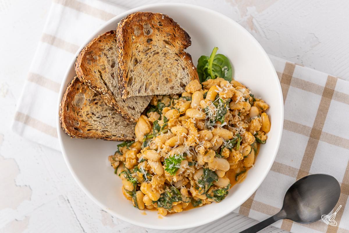 A bowl of Creamy White Bean and Spinach Sausage Skillet with sliced of toasted bread.